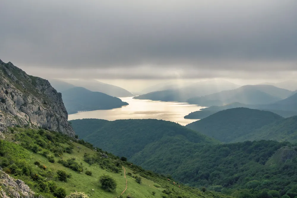 The Riaño reservoir seen from a grassy slope, a long stretch of water between wooded ridges, with sunlight breaking through low cloud.